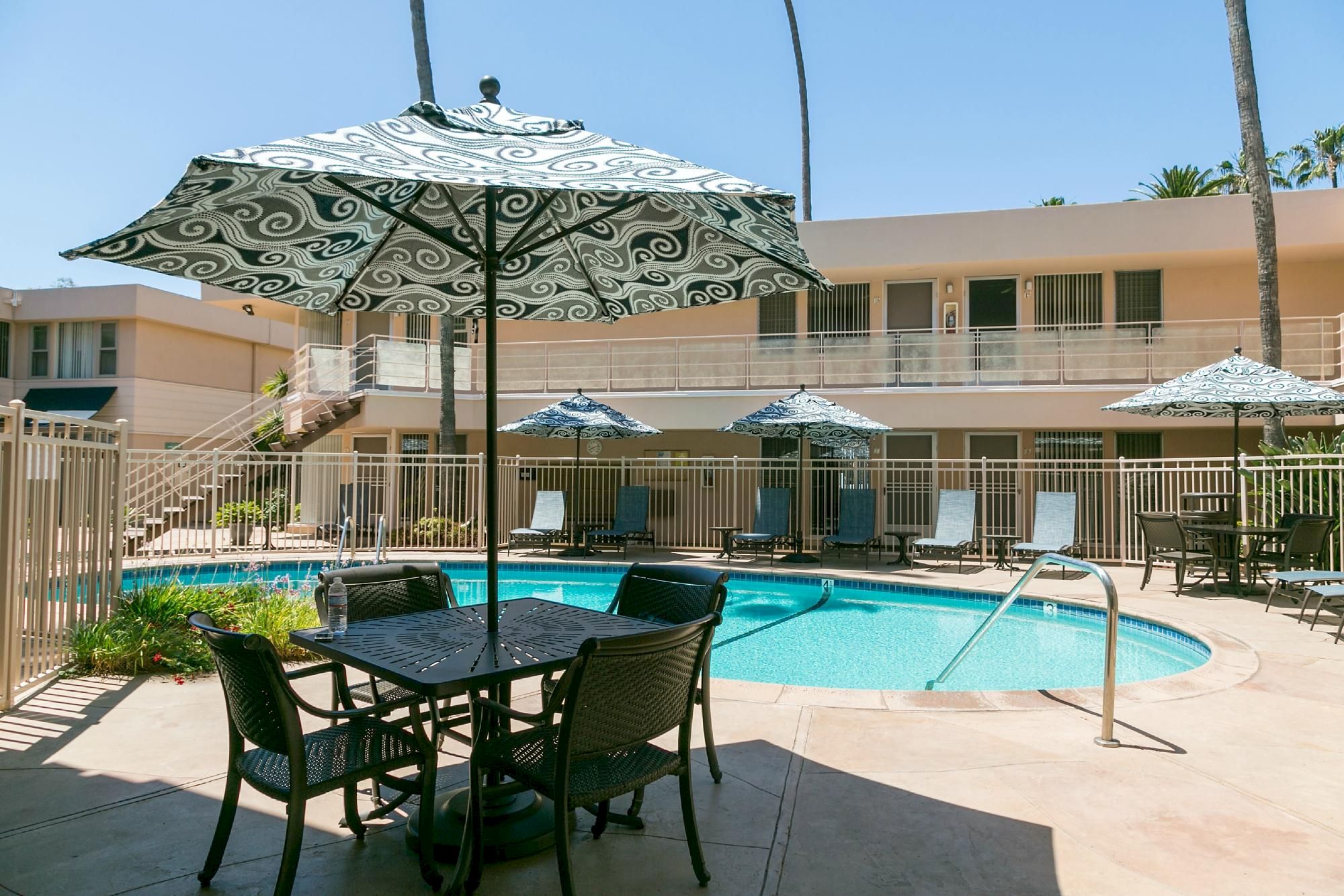 A sunny apartment complex pool area with a curved pool, patio tables, and striped umbrellas, surrounded by a beige building with balconies.