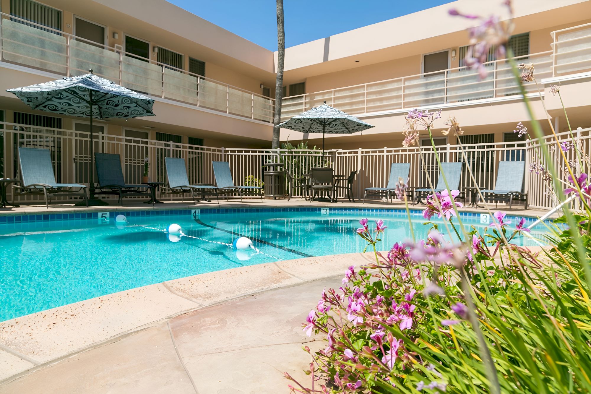 A hotel pool area with blue water, lounge chairs, umbrellas, and a courtyard, surrounded by a beige building with balconies.