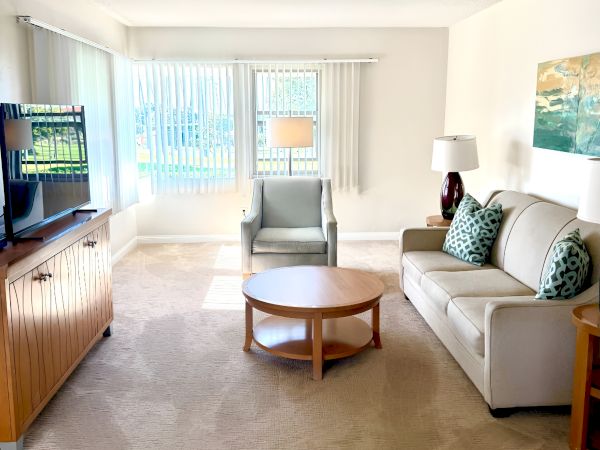 A bright living room with a beige carpet, white walls, a sofa with teal and beige cushions, a round coffee table, an armchair by a large window, and a TV on a wooden stand.