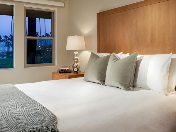 A neatly made hotel-style bedroom with a large bed, white linens, gray throw, plush pillows, wooden headboard, and matching nightstands with lamps.