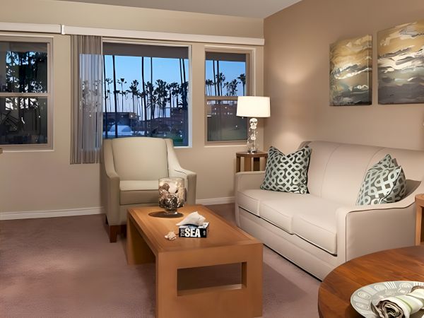 A cozy living room with beige walls, white sofa, patterned throw pillows, two chairs, wooden coffee table, lamps, and tropical wall art.