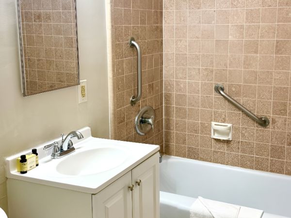 A small bathroom with a white vanity and sink, mirror, and light above; a tiled tub/shower with grab bars and a towel draped over the tub edge. End.