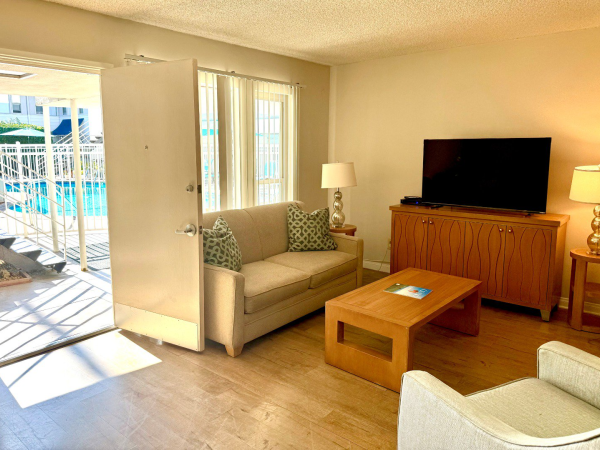 Cozy living room with a beige sofa, wooden coffee table, TV on a wooden console, two lamps, and sliding door leading to a sunny patio.