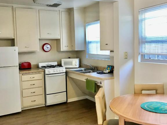 A small kitchen with white cabinets, a stove, fridge, window, ceiling fan, and a round dining table with chairs in a cozy, simple apartment.