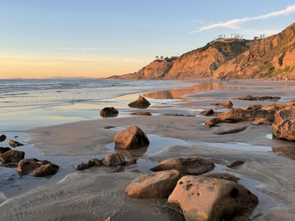 Rocky beach at low tide with wet sand, scattered stones, gentle waves, and a cliff-top path or houses in the distance at sunset/sunrise.