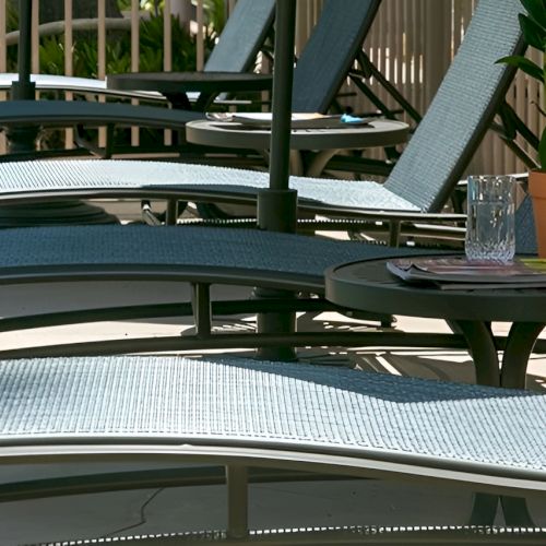 A row of empty poolside lounge chairs with small side tables, a plant in a pot, and a glass on one table, sunny outdoor setting.