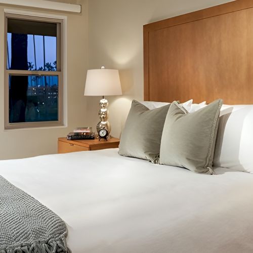 A tidy hotel room with a large bed, white linens, gray pillows, wooden headboard, and matching nightstands with lamps on each side.