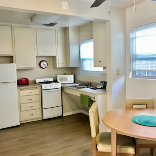 A small kitchen with white cabinets, a white stove, fridge, microwave, dining table and chairs, ceiling fan, and a windowed wall, cozy and simple.