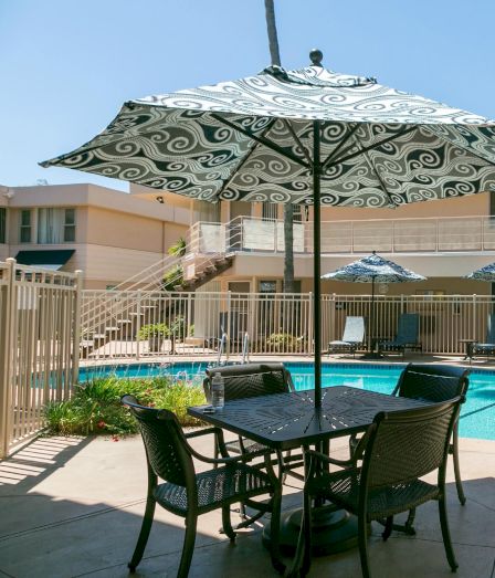 A sunny hotel pool area with a metal table and chairs under patterned umbrellas, surrounded by lounge chairs and a two-story building.