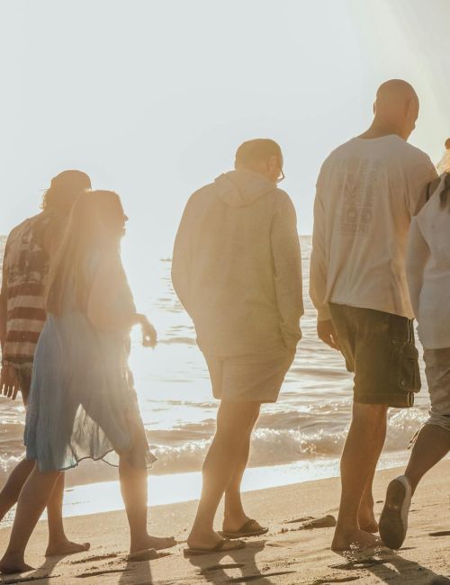 A group of six people walk along a sunny beach at sunset, casting long shadows on the sand.